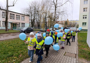 Dzieci ubrane w kamizelki odblaskowe spacerują uliczkami osiedla trzymając niebieskie balony