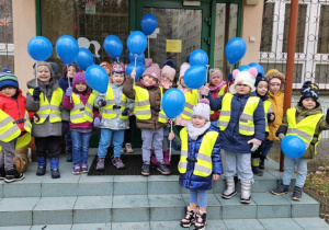 Dzieci ubrane w kamizelki odblaskowe pozują do zdjęcia na schodach przedszkola trzymając niebieskie balony