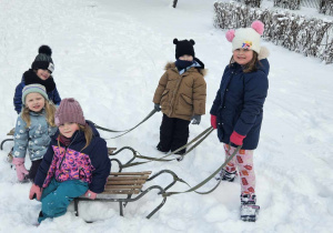 Zabawy w ogrodzie przedszkolnym - dwoje dzieci siedzi na sankach, dwoje dzieci ciągnie sanki, jedno dziecko kuca na śniegu