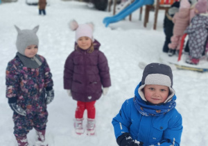 Troje dzieci stoi na śniegu w ogrodzie przedszkolnym.