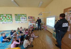 Dzieci siedzą na dywanie i uczestniczą w zajęciach prowadzonych przez inspektorów ITD