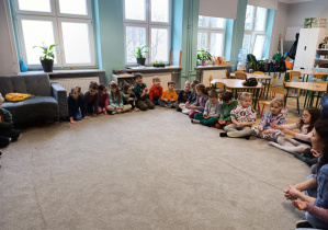 Dzieci siedzą na dywanie i uczestniczą w zajęciach w szkole podstawowej