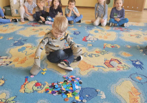 Dzień Świadomości Autyzmu - dzieci siedzą w kole, chłopiec w środku koła próbuje chwycić klocki za pomocą rękawic kuchennych