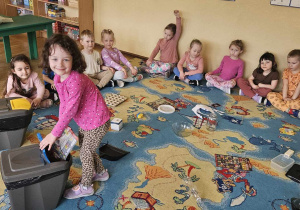 Dbamy o przyrodę - dzieci siedzą w kole, w środku rozsypane są śmieci, dziewczynka wrzuca gazetę do odpowiedniego kosza na śmieci
