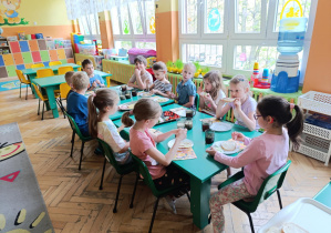 Dzieci siedzą przy stolikach jedząc śniadanie wielkanocne