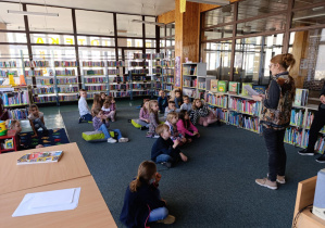 Dzieci siedzą na dywanie w bibliotece słuchając bajki czytanej przez panią bibliotekarkę