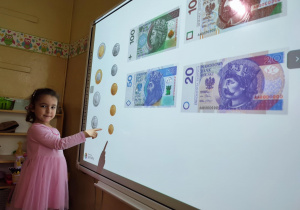 Zajęcia o pieniądzach - dziewczynka stoi obok tablicy multimedialnej i wskazuje odpowiednią monetę
