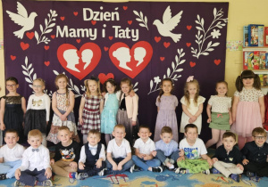Dzień Mamy i Taty - dziewczynki stoją, chłopcy siedzą przez dziewczynkami na tle dekoracji