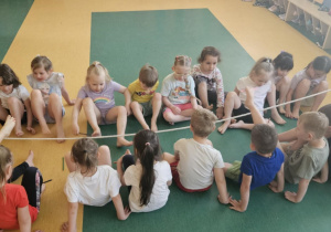 Gimnastyka - dzieci na sali gimnastycznej siedzą w dwóch rzędach na przeciwko siebie przy linie i przekładają kredki na stronę przeciwnika za pomocą stóp