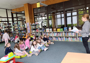 Dzieci siedzą na dywanie i słuchają książki czytanej przez panią bibliotekarkę