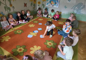 Dzieci siedzą w kole na dywanie, dziewczynka dopasowuje kolorystycznie kropki do słoików.