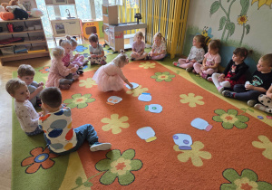 Dzieci siedzą w kole na dywanie, dziewczynka dopasowuje kolorystycznie kropki do słoików.