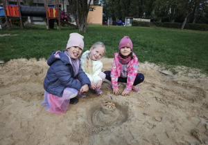 Trzy dziewczynki kucają w piaskownicy przy swojej budowli z piasku