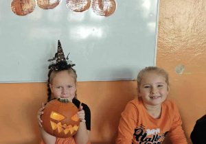 Dwie dziewczynki siedzą na dywanie i trzymają swoje ozdobione dynie