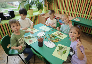 Dzieci siedzące przy stole przylejaja liście na jesiennej pracy plastycznej