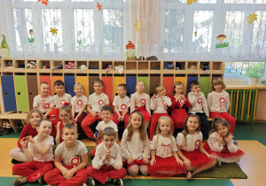 Dzieci ubrane na biało czerwono siedzą na ławce i na podłodze