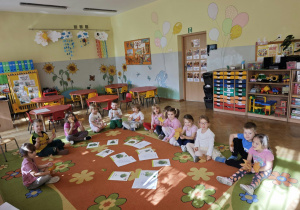 Dzieci siedzą w półkolu na dywanie obok ilustracji przedstawiających drzewa