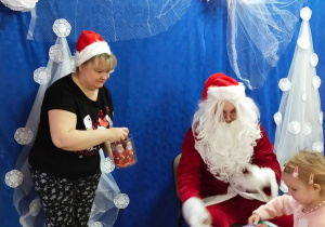 Mikołaj podający dziewczynce prezent i pani w mikołajowej czapeczce szykuje się podać czekoladkę