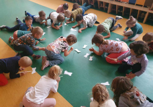 Dzień Kundelka - dzieci siedzą na podłodze na sali gimnastycznej i dmuchają za pomocą słomek w kostki wykonane z papieru