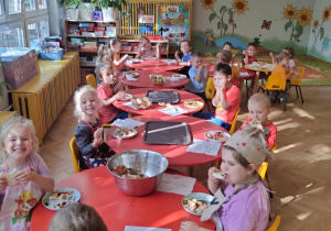 Dzieci siedzą przy długim stole i jedzą wykonaną przez siebie salatkę owocową