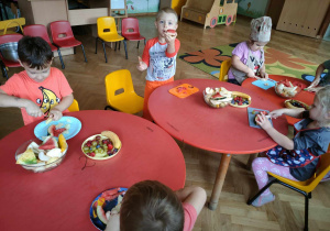 Dzieci siedzą przy stolikach i kroją owoce na salatkę owocową