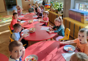 Dzieci siedzą przy długim stole i wspólnie jedzą sałatkę owocową wykonaną przez siebie