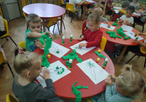 Dzieci siedzą przy stolikach i wykonują z zielonej bibuły kulki do liści marchwi