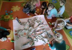 Zabawy ze sztuką - dzieci siedzą na dywanie i doklejają kółeczka do drzewa wykonanego z gazet