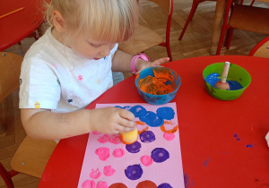 Dziewczynka wykonuje pracę plastyczną ,,Kolorowe kropki", stemplując gąbką zamoczoną w farbie po kartce