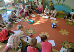 Dzieci siedzą na dywanie i układają na kartonikach tyle kasztanów ile jest na kostce oczek