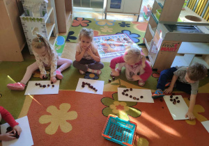 Dzieci siedzą na dywanie i układają obrazki z kasztanów na kartonach