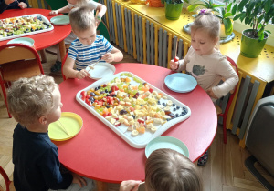 Dzieci siedzą przy stoliku i zaczynają nabijać kawałki owoców na wykałaczki
