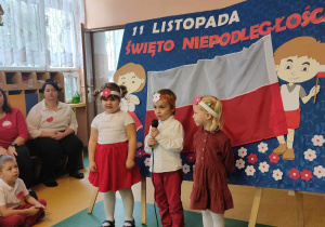 Święto Niepodległości - dwie dziewczynki i chłopiec trzymający mikrofon na tle dekoracji