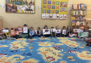Dzieci siedzący na dywanie pokazują swoje prace plastyczne