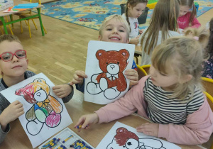 Dzieci siedzące przy stole pokazują swoje rysunki misiów