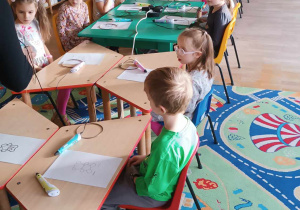 Dzieci siedzą przy stolikach ustawionych w rzędzie i uczestniczą w warsztatach z długopisami 3D