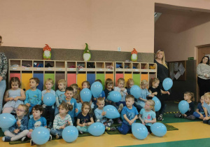 Dzieci siedzące na ławeczkach i podłodze ubrane na niebiesko trzymają niebieskie balony