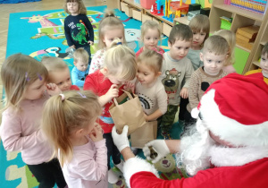 Dzieci stoją wokół Mikołaja, a dziewczynka zagląda do swojej paczki z prezentem