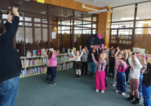 Zajęcia w bibliotece - dzieci wraz z bibliotekarzem podnoszą ręce do góry