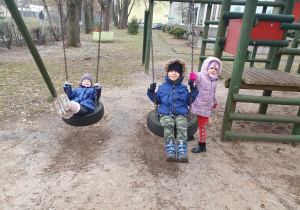 Zabawy w ogrodzie przedszkolnym - dwoje dzieci huśta się na huśtawkach, jedno z nich stoi obok