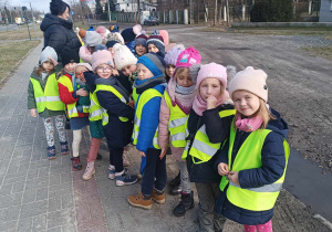 Grupa dzieci ubrana w kamizelki odblaskowe spaceruje uliczkami osiedla
