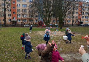 Dzieci będąc w ogrodzie łapią mydlane bańki puszczane przez nauczyciela