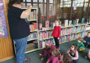 Biblioteka - dzieci siedzą na dywanie jedno dziecko stoi obok bibliotekarza i odgaduje zagadkę