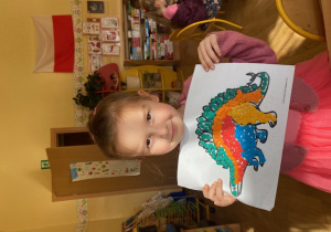dziewczynka prezentuje pracę plastyczną przedstawiającą kolorowego dinozaura