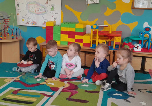 Dzieci siedzą przed kolorowymi krążkami, na których położone są białe, papierowe motyle