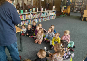 Dzieci siedzą na dywanie w bibliotece i rozmawiają z bibliotekarzem na temat miejsca, w którym się znajdują