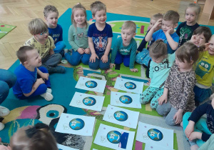 Dzieci siedzą na dywanie w kole; na środku rozłożone są ilustracje planety- Ziemi i zadania do wykonania