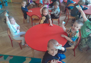 Dzieci siedząc przy stoliku pokazują wykonane przez siebie grzechotki z rolek po papierze