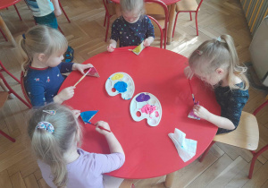 Dzieci siedząc przy stolikach malują farbami swoje grzechotki