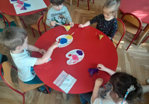 Dzieci siedząc przy stolikach malują farbami swoje grzechotki